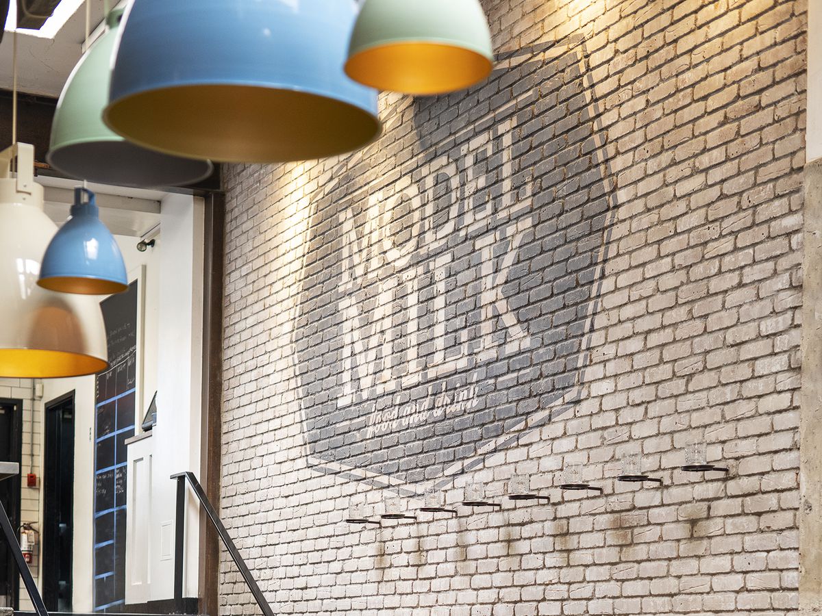 An interior brick wall decorated with the logo for Model Milk, behind eggshell blue pendant lights hanging over a long wooden table set for a meal
