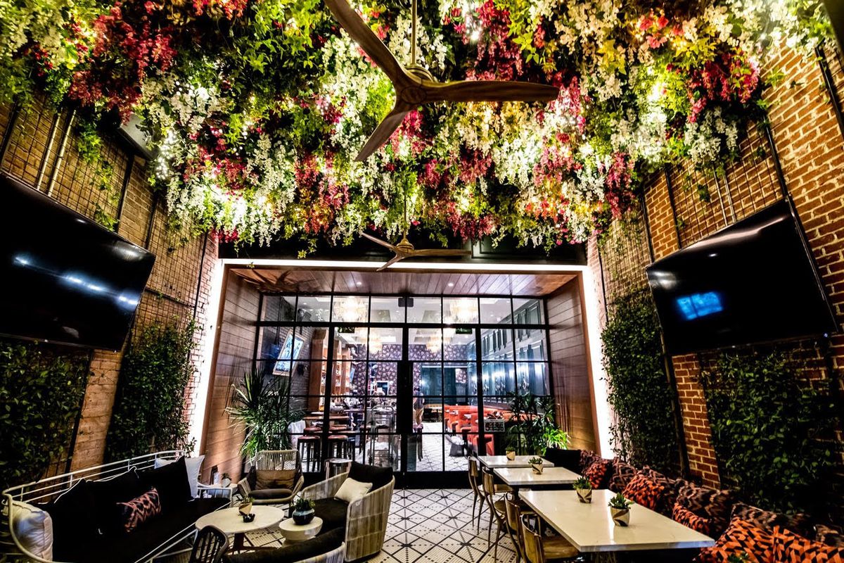 A patio space with lush greenery hanging from the ceiling, glassed doors, and tables with chairs 