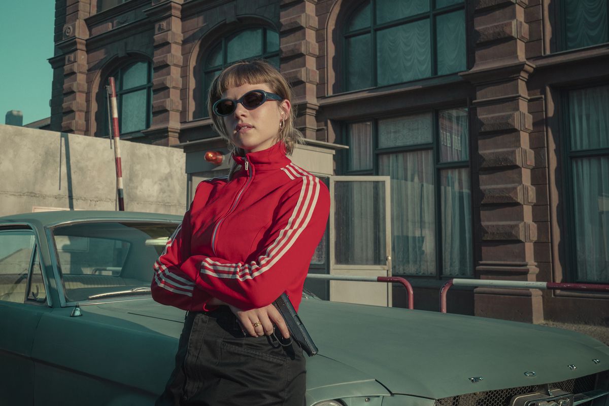 Jelia Haase as Kleo wears a red tracksuit and jeans and holds a pistol while leaning against a car. She also is wearing sunglasses and looks cool as hell.