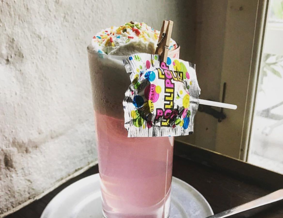A pink cocktail in a highball glass topped with whipped cream and sprinkles, and a lollipop clothespinned to the edge of the glass, sitting on a small dish with a long ice cream spoon, all against a corner of a bar against a cement wall