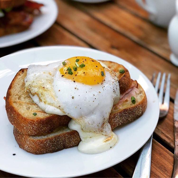 A very runny egg topped croque madame, on a plate beside a fork on a wooden patio table.