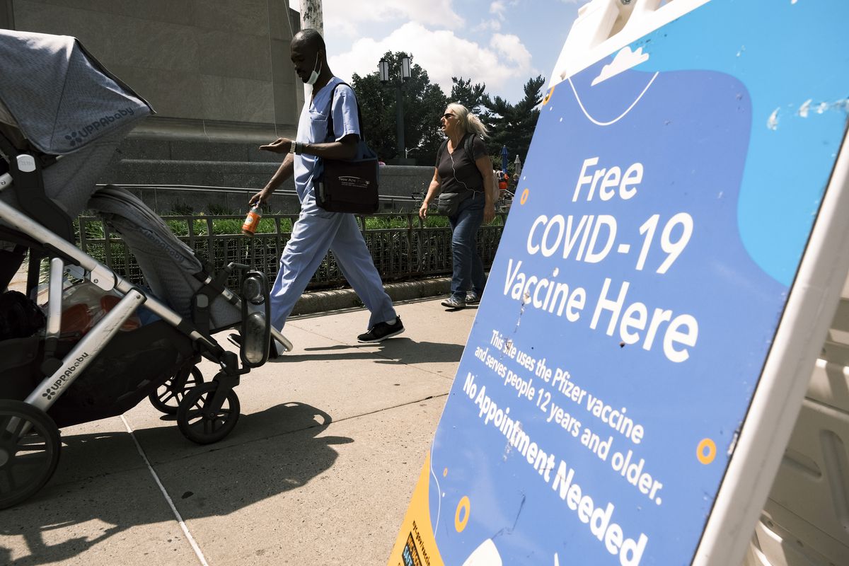 Pedestrians, one pushing a stroller, walk past a sidewalk sign that reads, “Free Covid-19 vaccine here.”