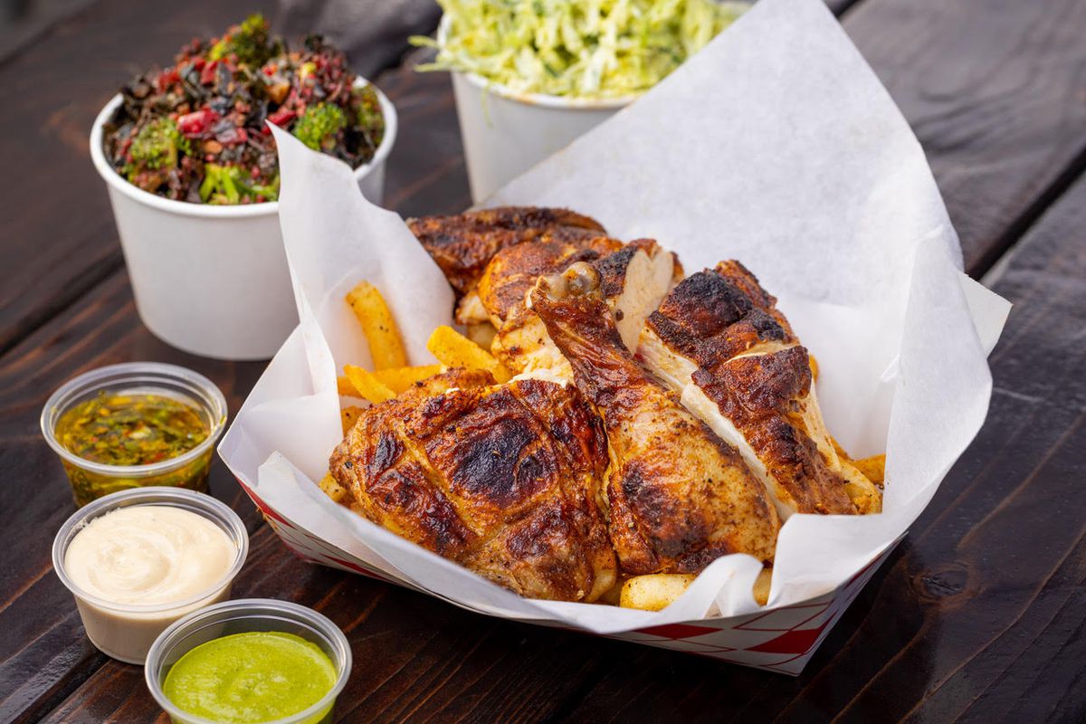 Crispy-skinned chicken over fries with sauces in plastic containers, on a wooden table.