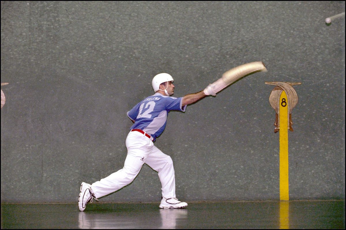 A jai alai player in white pants and blue jersey, reaches out with a hook-shaped basket on his right arm as he tries to catch a ball.