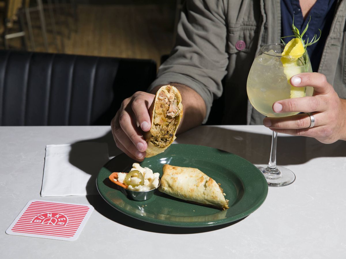 A fan digging into a burrito that cut in half on a green plated with another hand holding a wine glass with a yellow colored drink.