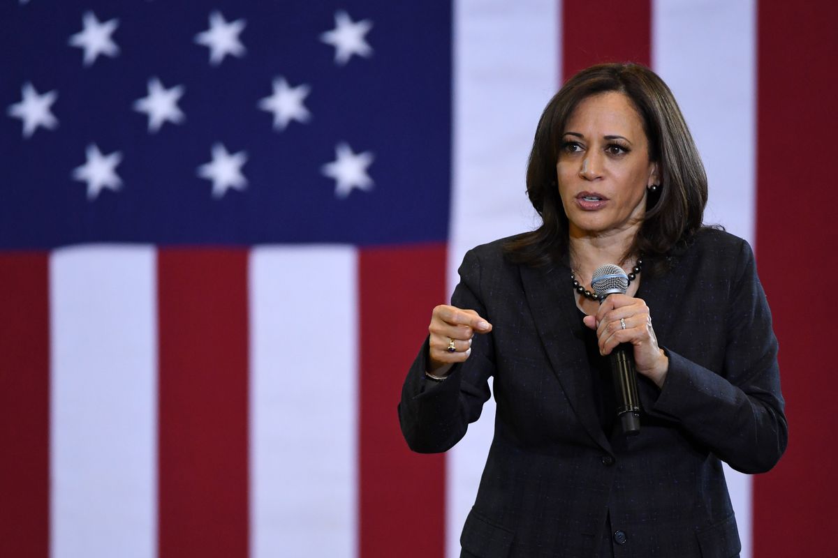 Harris, in a black suit, speaks in front of a giant American flag.