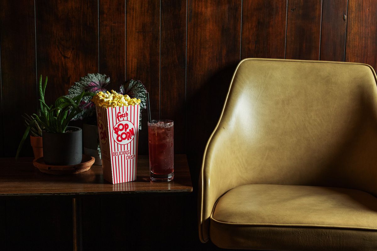 A red and white striped box of buttery popcorn and a tall cocktail with a black straw sit on a mid-century table next to an antique pleather chair.