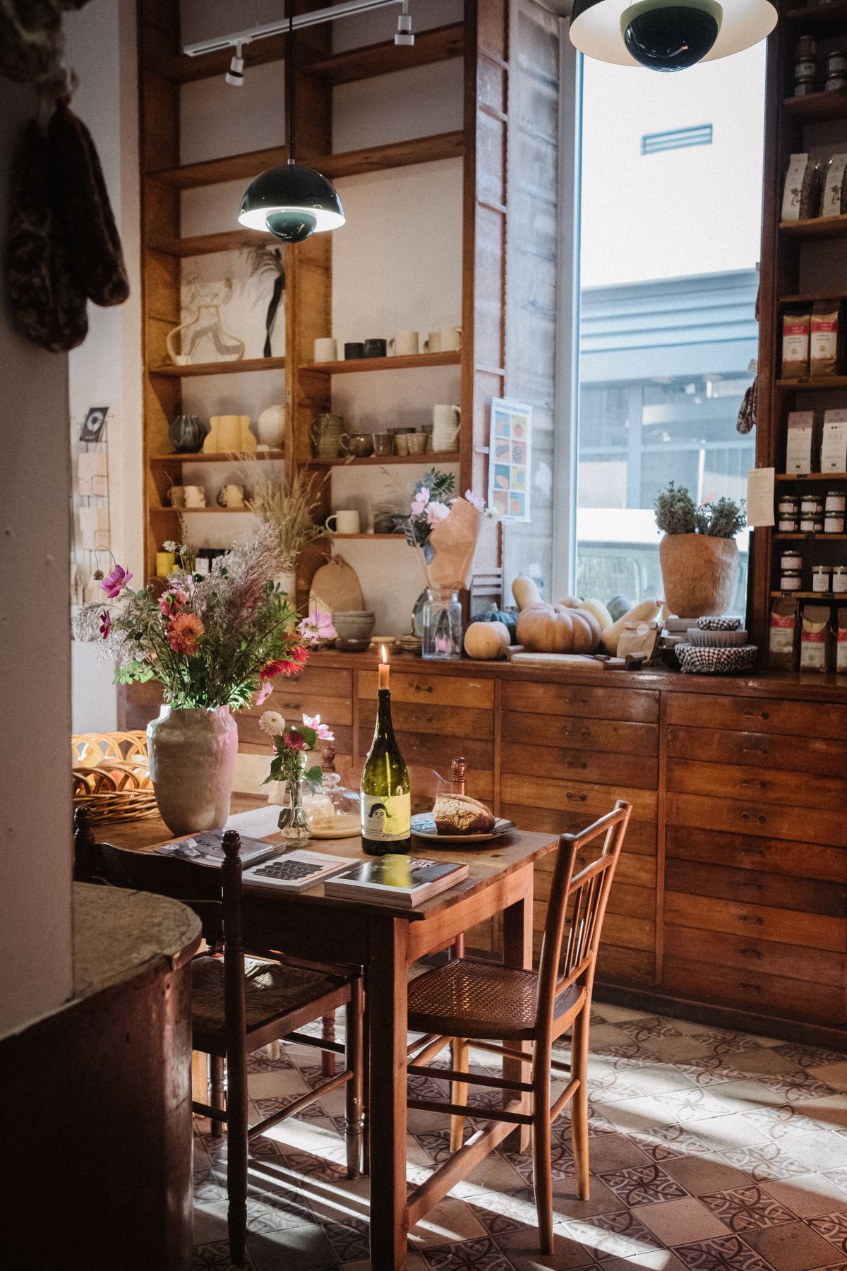 A candle burns in an empty wine bottle, sitting on a table beside a pot of flowers and food books. Beyond is a sun-lit interior, with shelves of other items and dark wood drawers.