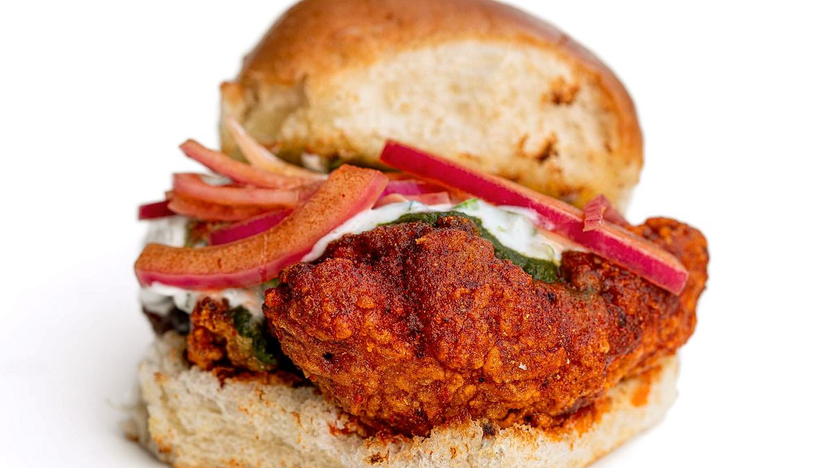 A slider-sized fried chicken sandwich with white and green sauces plus pickled onions, on a small toasted bun.