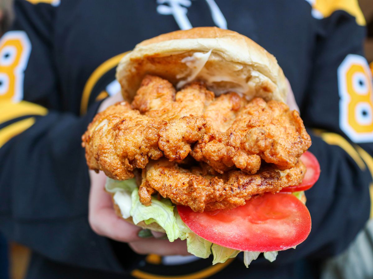 A person in a Bruins jersey holds out a fried chicken sandwich in front of their body.