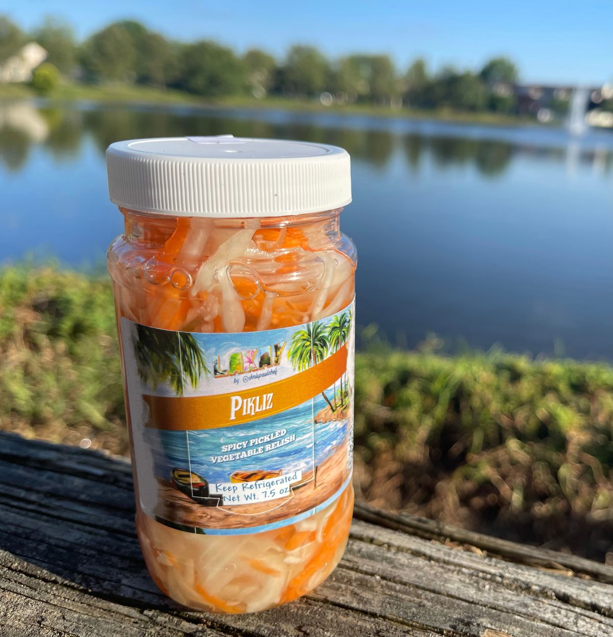 A jar of Lakay Pikliz pickled vegetables with a white lid on a bench looking out over a lake.