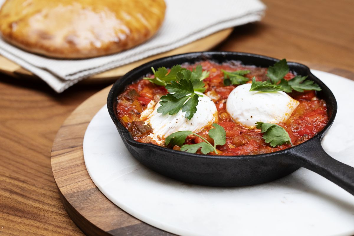 Shakshouka in a cast iron dish on a white plate on a wooden table, with bread visible in the background. 