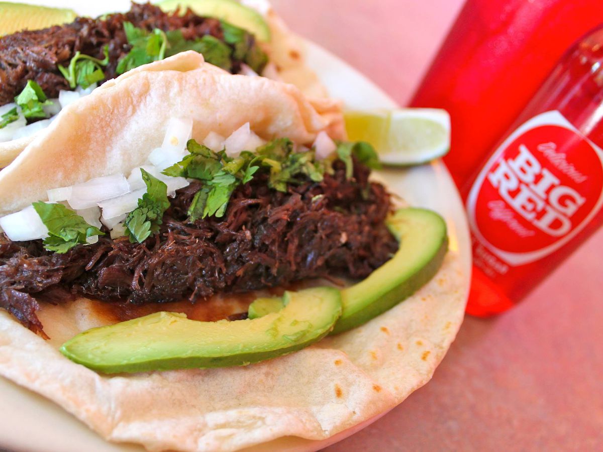 A closeup of two barbacoa tacos with avocado and onions next to a glass bottle of Big Red soda.
