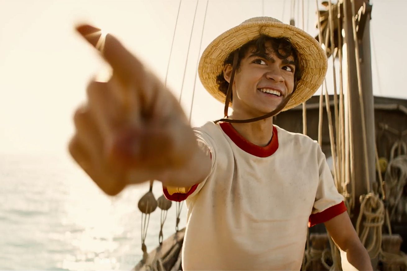 A boy wearing a straw hat and a white t-shirt ringed with red around the collar and sleeves. The boy is smiling, pointing towards the camera, and standing on the deck of a boat.