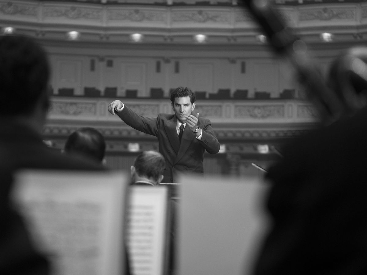 Bradley Cooper conducting an orchestra seen from the middle row in a black-and-white scene from Netflix’s Maestro