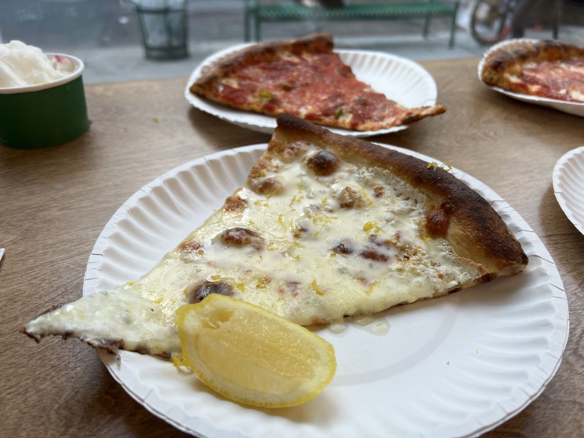 A white slice of pizza, sprinkled with lemon zest, sits on a paper plate, with a lemon wedge on the site; cheese slices are visible in the background