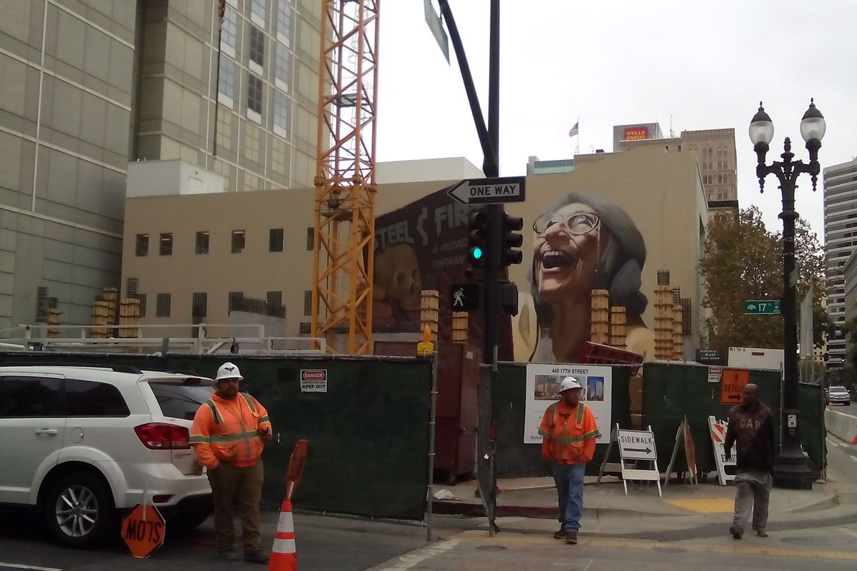 A construction site in front of a mural painted on the side of an Oakland building depicting an elderly woman listening to heavy metal music.