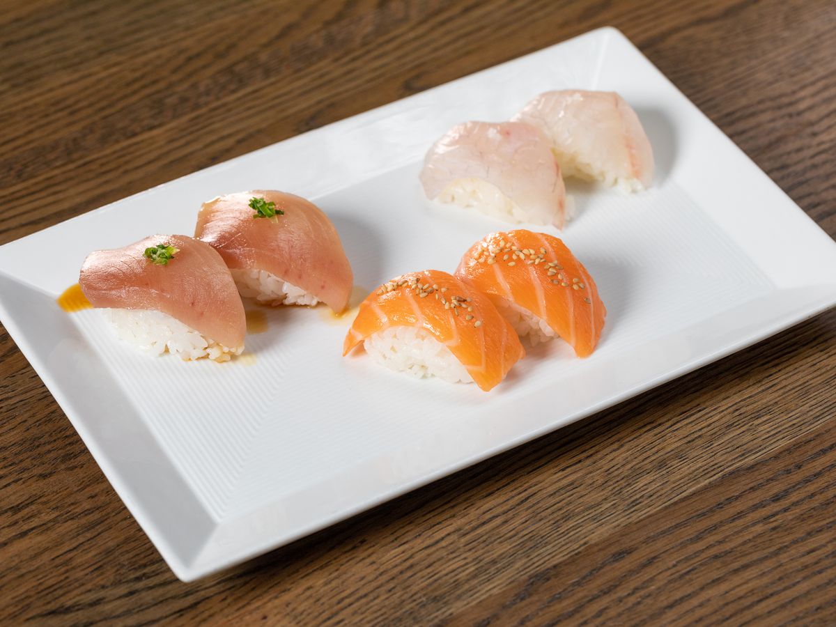Three pairs of two pieces of sushi, topped with raw cuts of colorful fish, sit on a rectangular plate