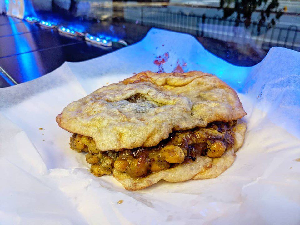 A serving of doubles —&nbsp;spicy and sweet chickpeas sandwiched between two pieces of fried dough —&nbsp;sits on white paper on a counter, illuminated by a glowing blue light