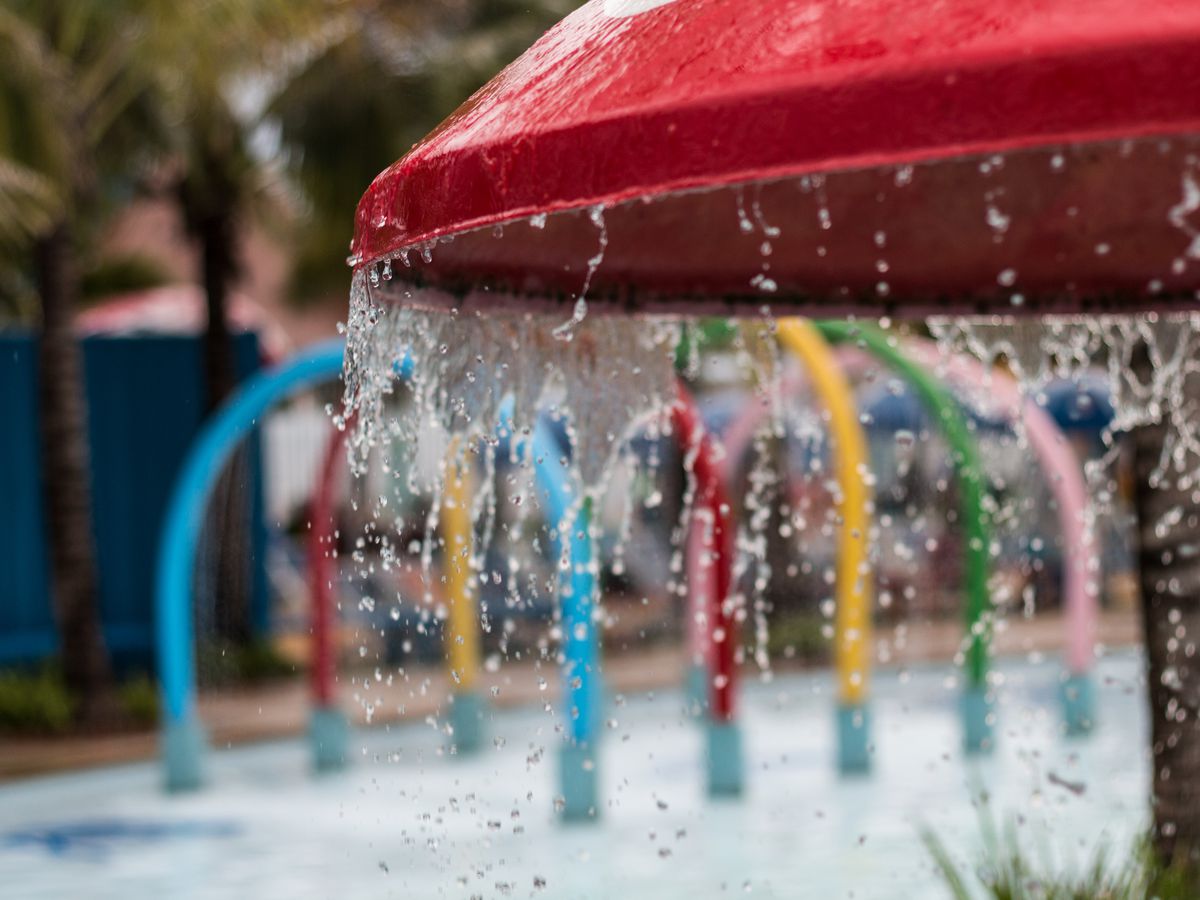 A red mushroom-shaped structure dips water at a spray park, with multicolor sprinklers in the background.