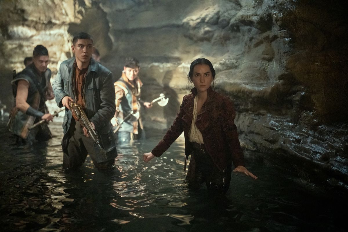 From left to right, Tolya (Lewis Tan), Mal (Archie Renaux), Tamar (Anna Leong Brophy), and Alina (Jessie Mei Li) stand knee deep in a water-filled cave in Shadow and Bone season 2. Toylya holds a sword, Tamar an axe, and Mal a gun, while Alina’s hands are bar her side signaling them to hold their places.