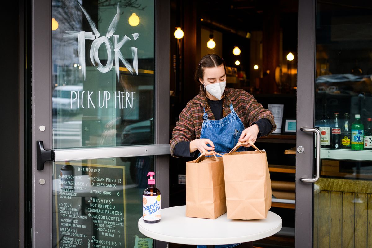 A woman with braids, a mask, and a denim apron brings two paper bags to a small white table in the door of Toki. A bottle of hand sanitizer sits on the table.