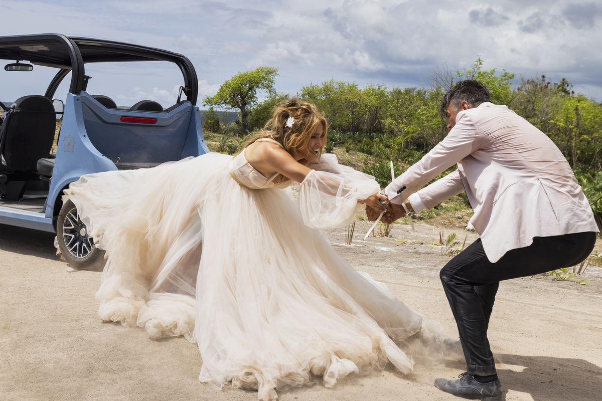 Josh Duhamel holds on to Jennifer Lopez’s arms as she stretches out from the back of a car in Shotgun Wedding. Both are wearing wedding attire.