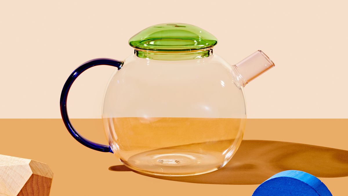 A round shaped clear glass teapot with a clear green lid and a thin, geometric dark blue handle.