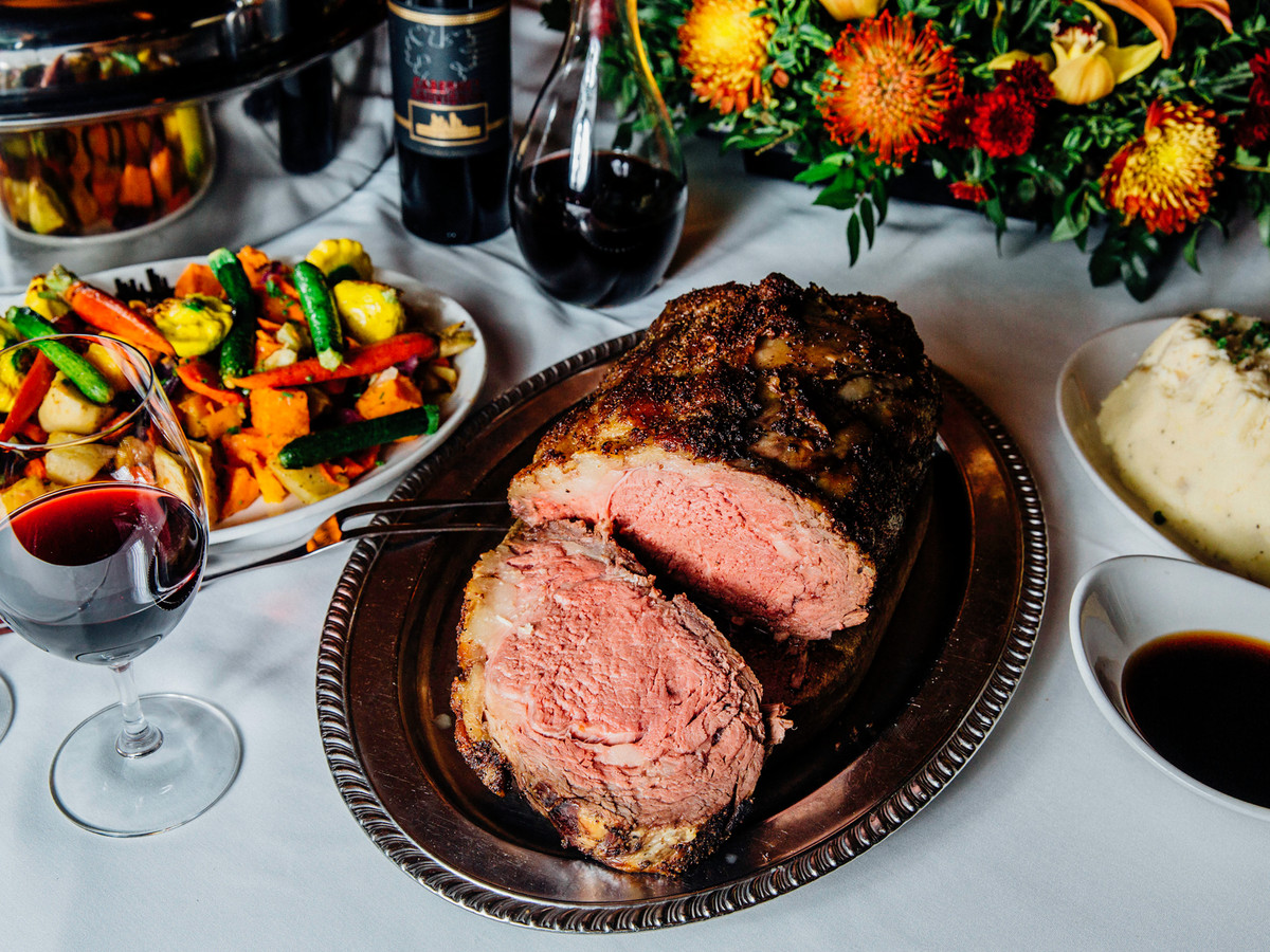A feast spread out on a white tablecloth. A slice prime rib on a silver platter is front and center. Mashed potatoes, flowers, and a wine glass are nearby.