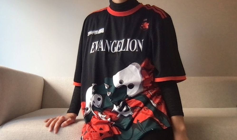 a person sits on a couch wearing a b lack and red evangelion jersey 