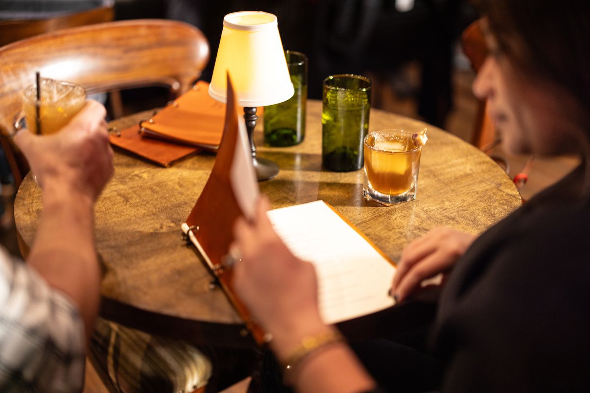 Two people are sitting at a circular table. One holds a cocktail in hand. The other is looking at a menu.