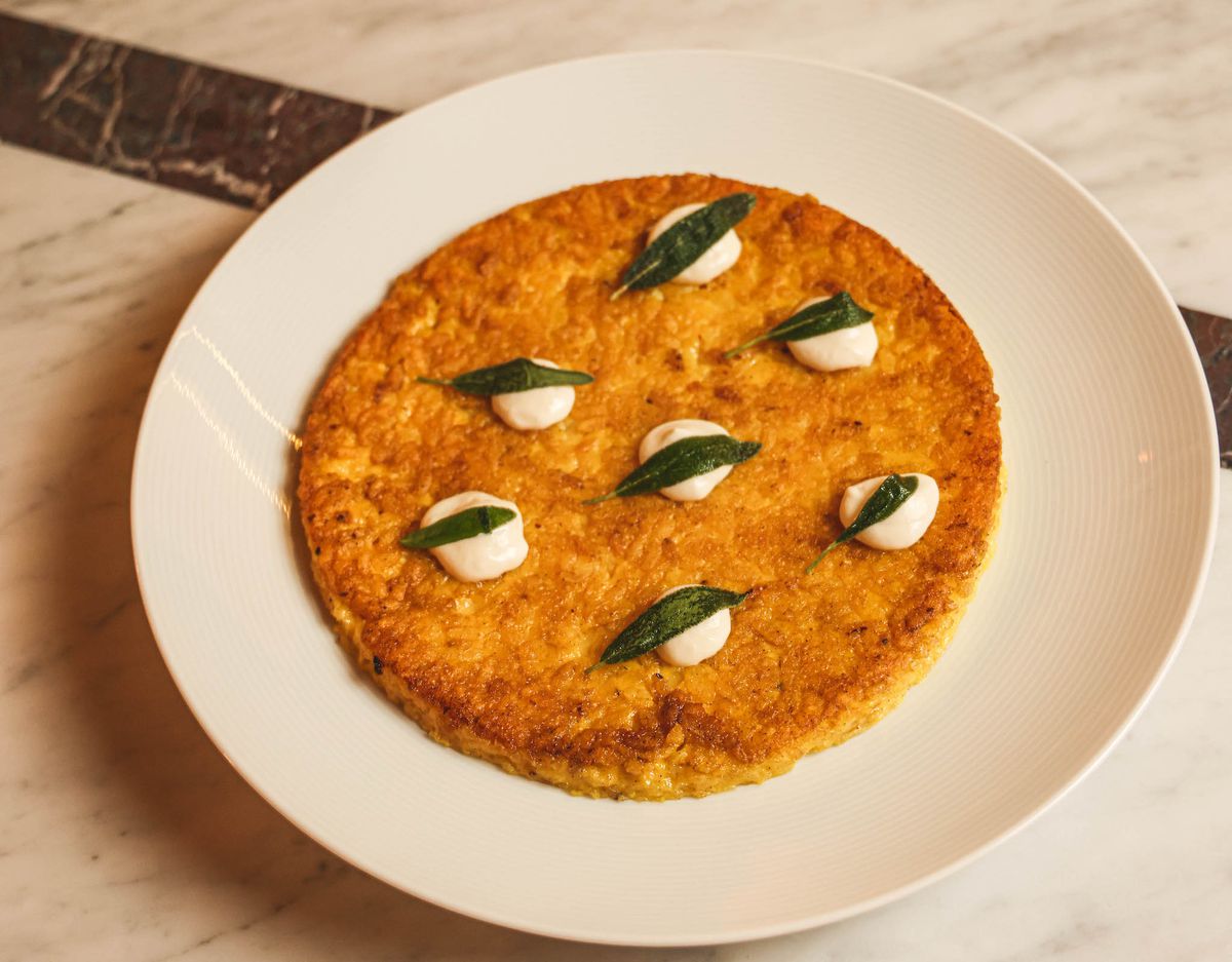 A white plate with a saffron risotto pancake with dotes of cream and sage.