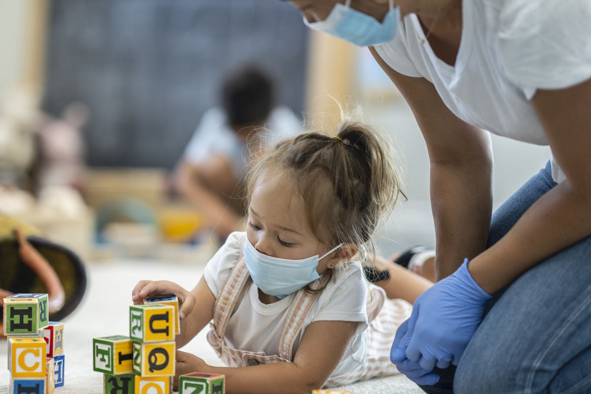 Young girl in mask plays with blocks while masked and gloved woman watches