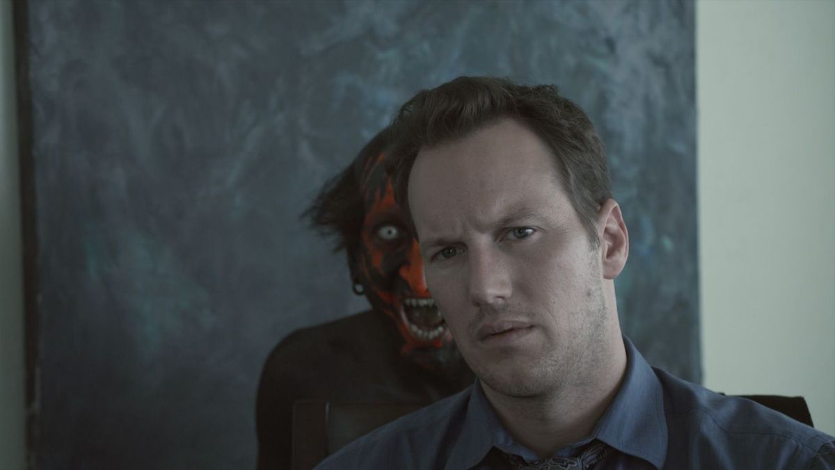 Un demonio de cara roja frunce el ceño burlonamente por encima del hombro de Josh Lambert (Patrick Wilson) en Insidious de James Wan.