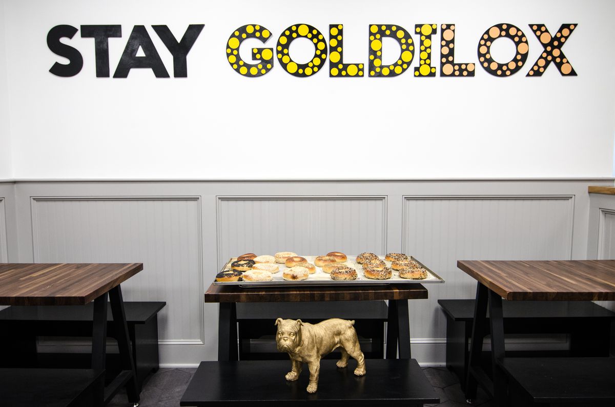 The seating area at bagel shop Goldilox Bagels in Medford. Lettering on the wall reads “Stay Goldilox.” A tray of bagels sits on a table, and a golden bulldog statue sits on a bench in front of the table.