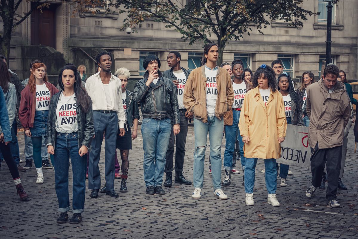 It’s a Sin cast protests on a cobblestone road in “Aids needs Aid” t-shirts