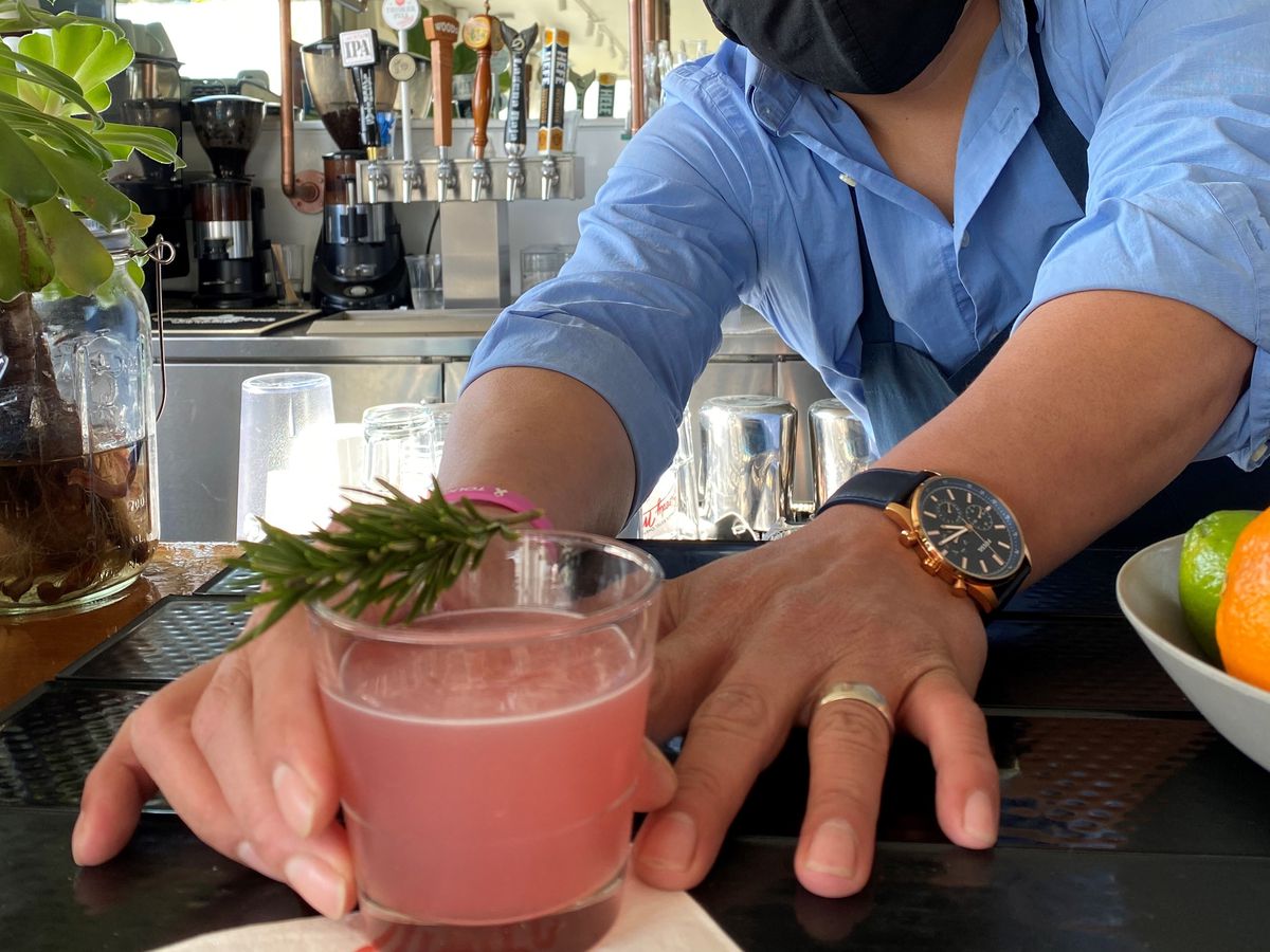 A masked bartender hands over a light pink cocktail garnished with a rosemary sprig laying across the top of the rim.
