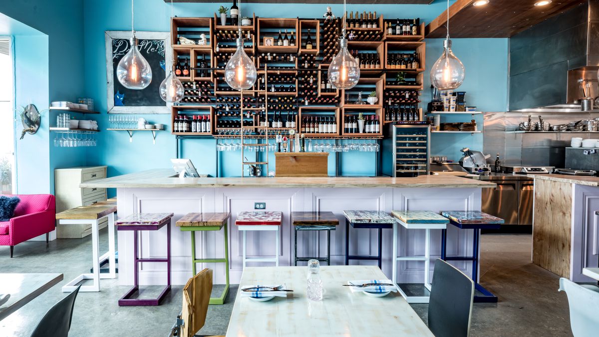 A restaurant dining room with a teal-blue wall and wine bottles on shelves and a bar and a dining table.