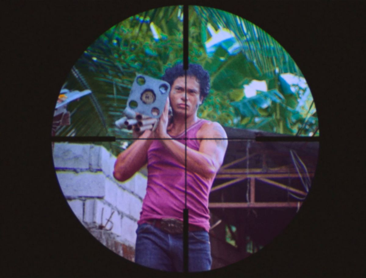 A Filipino man with curly hair, wearing a pink muscle shirt, is seen in low-res video through a sniper’s sight in a movie-within-a-movie sequence from Leonor Will Never Die