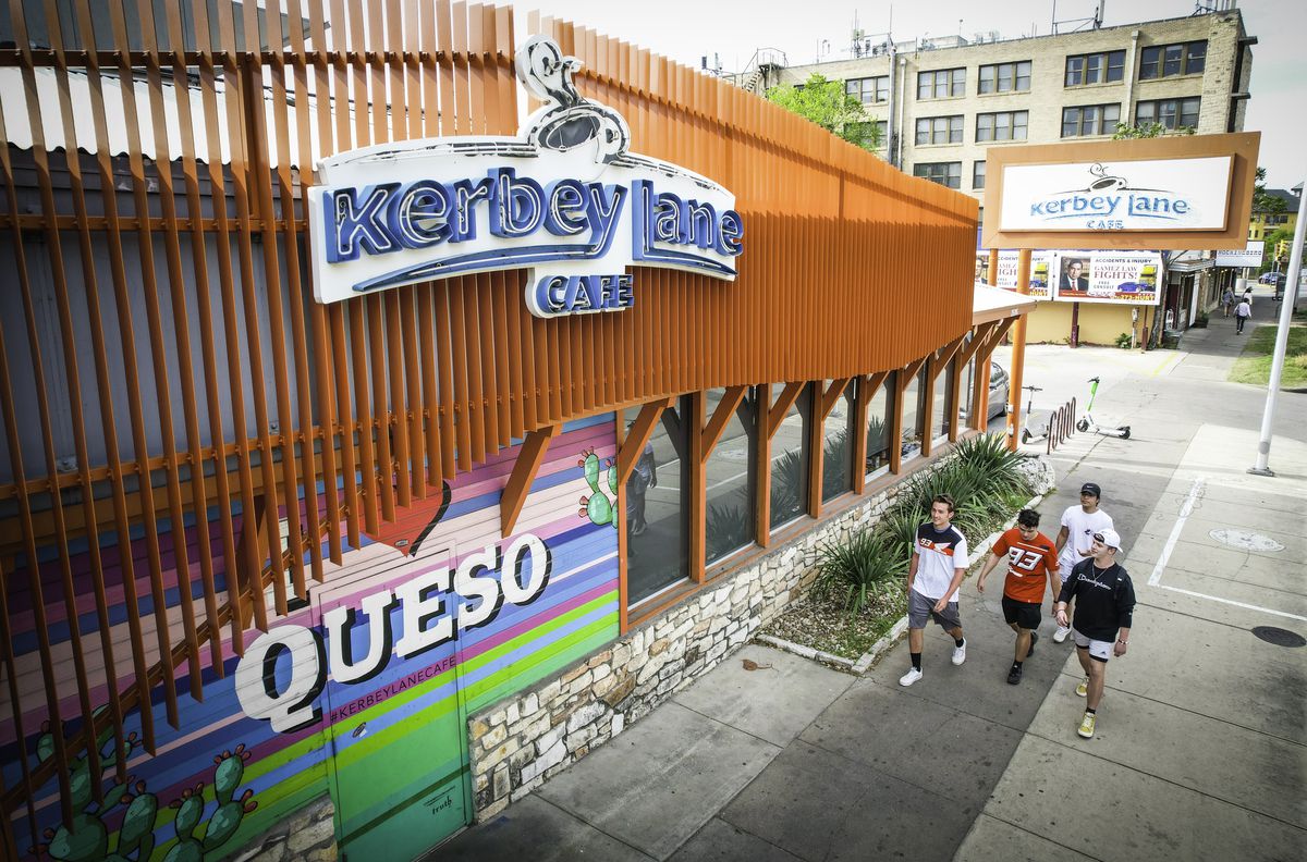 A restaurant facade with vertical red panels and a blue neon sign reading “Kerbey Lane Cafe” and a colorful wall with the words “Queso” on it and a group of four people walking by on the sidewalk. 