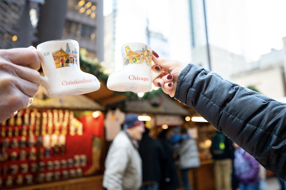 Two people holding small boot-shaped mugs cheers outside a Germanic-looking Christmas market stall.
