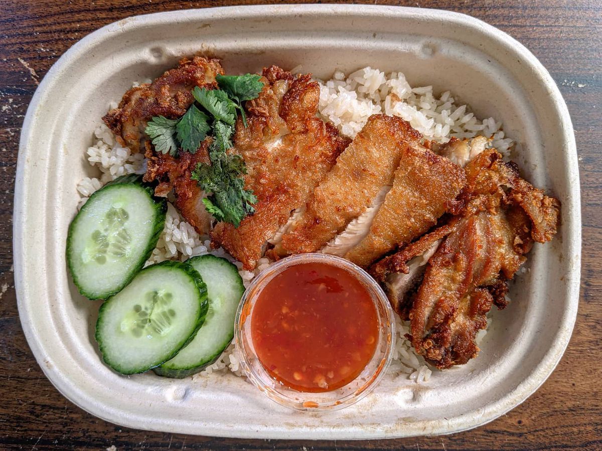 overhead view of a compostable takeout container of crispy chicken slices over rice, with cucumber and cilantro garnish and a plastic cup of a sweet chile sauce