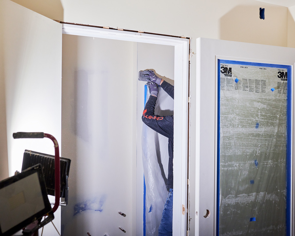 A worker sands and paints the interior of a tiny house being built, using blue tape and white paint.