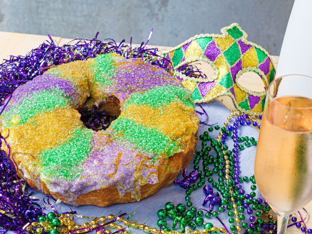 A King Cake decorated with cream cheese and different colored sparkles sits next to a mask and a glass of champagne at Eunice.