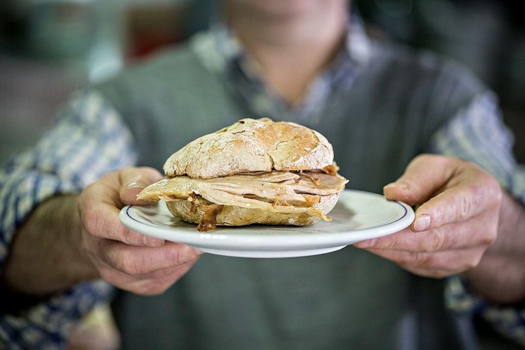 A man holds out a plate with both hands bearing a pork sandwich on thick crusty bread