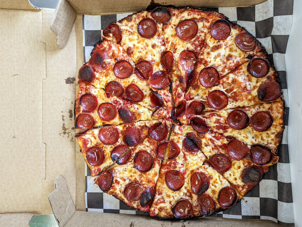 Overhead view of a pepperoni pizza on black and white checkered paper in a pizza box. The pizza has a charred edge with crust barely visible.