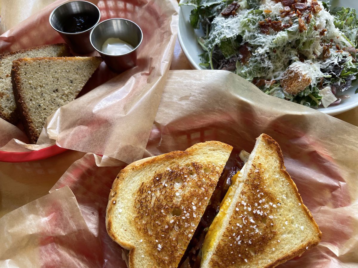 From above, a sliced grilled cheese in a paper-lined basket, beside a salad and another bread-based dish. 