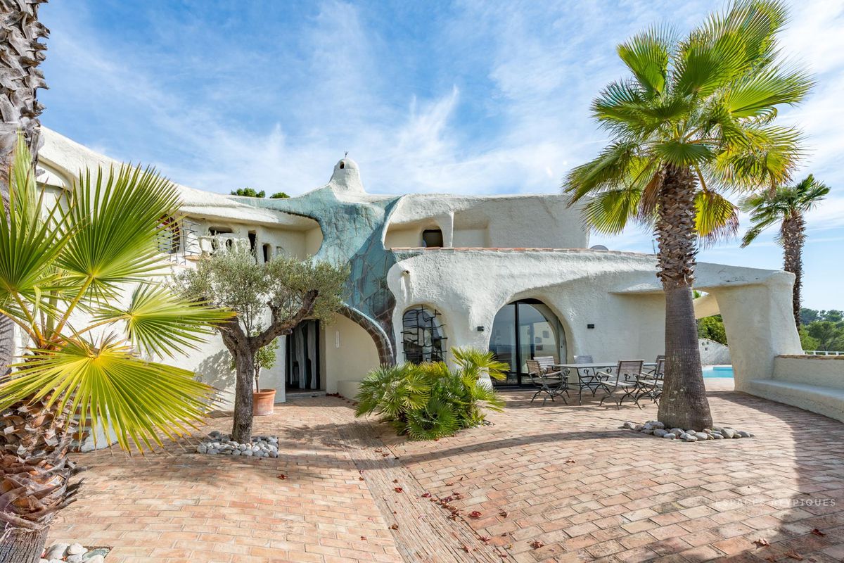 Shot of structure made from rough materials feature curved walls and windows with brick courtyard and palm trees surrounding it. 