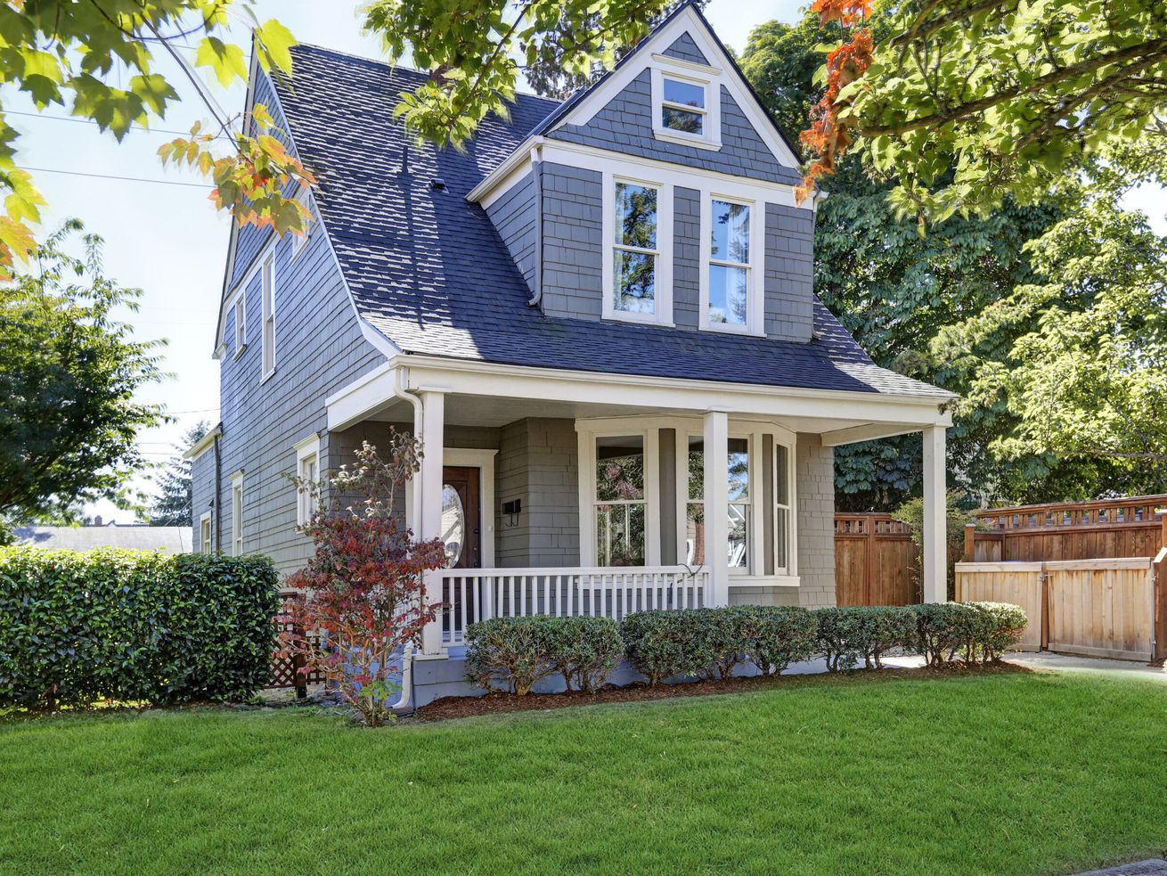A grey-blue home with a large front porch, large green front yard, and several green shrubs.