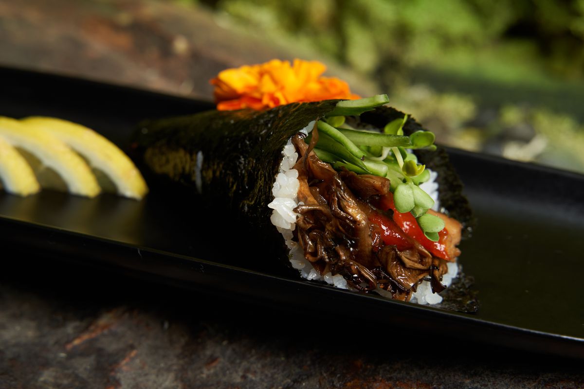 A close up of a Japanese handroll with mushrooms on a slate plate with orange flower on top.